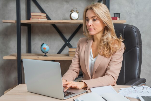 Retrato de una empresaria joven que se sienta en el lugar de trabajo usando el ordenador portátil en oficina