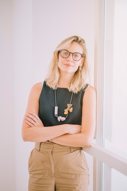 Retrato de una empresaria joven rubia confiada con los brazos cruzados que se inclinan en la pared de cristal