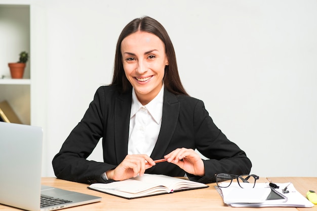 Retrato de una empresaria joven sonriente que se sienta en el escritorio con el lápiz y el diario