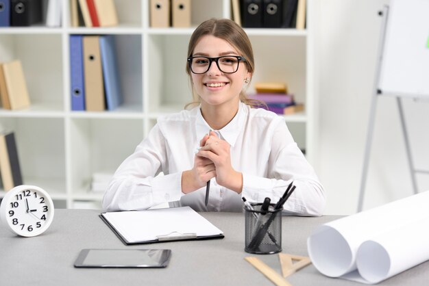 Retrato de una empresaria joven sonriente que se sienta en el lugar de trabajo con el tablero; despertador; soporte para celular y lapiz en escritorio