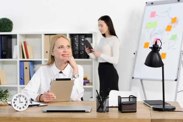 Retrato de una empresaria joven sonriente que sostiene la tableta digital que mira lejos con su colega que se coloca en el fondo