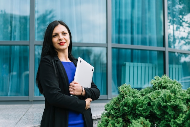 Retrato de una empresaria sonriente que sostiene el ordenador portátil delante del edificio de cristal