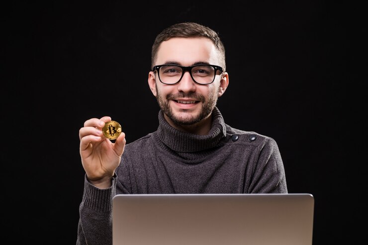Foto gratuito retrato de un empresario feliz mostrando bitcoin mientras está sentado en un escritorio con ordenador portátil aislado sobre negro