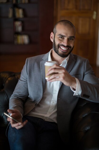 Retrato del empresario sonriente sosteniendo un teléfono móvil y una taza de café en la sala de espera