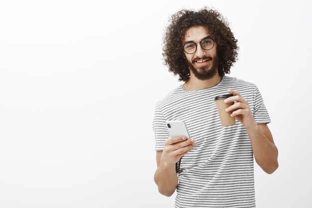 Retrato de encantador chico juguetón con barba y cabello rizado, tomando café y sosteniendo un teléfono inteligente, mirando con curiosidad