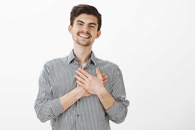 Retrato de encantador feliz hombre caucásico con barba y bigote, sosteniendo las palmas de las manos en el corazón y sonriendo de satisfacción, siendo tocado con gesto agradable