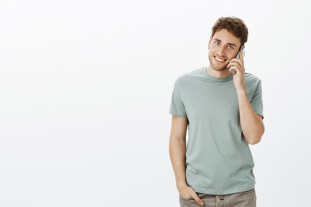 Retrato de encantador hombre rubio saliente con cerdas, sosteniendo la mano en el bolsillo mientras espera que mamá coja el teléfono, llamando y sosteniendo el teléfono inteligente cerca de la oreja