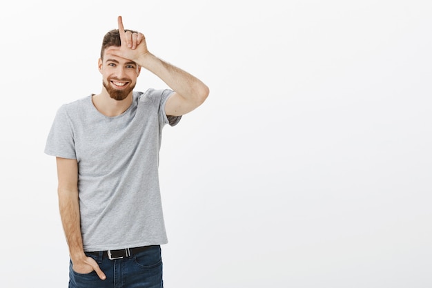 Retrato de encantador macho europeo juguetón y masculino con barba, ojos azules y bigote sosteniendo la mano en jeans mostrando la letra L sobre la frente como si se burlara de un amigo haciendo un gesto de perdedor y sonriendo