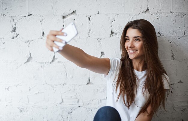 Retrato de encantador modelo femenino europeo, haciendo selfie en smartphone cerca de la pared de ladrillo blanco, sonriendo alegremente. La bloguera de moda toma una foto para publicarla en su blog. Tiene muchos fans.