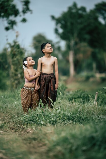 Retrato de un encantador niño asiático sin camisa y una niña en traje tradicional tailandés y poner una hermosa flor en su oreja, de pie de la mano y mirando al cielo con una sonrisa, espacio de copia