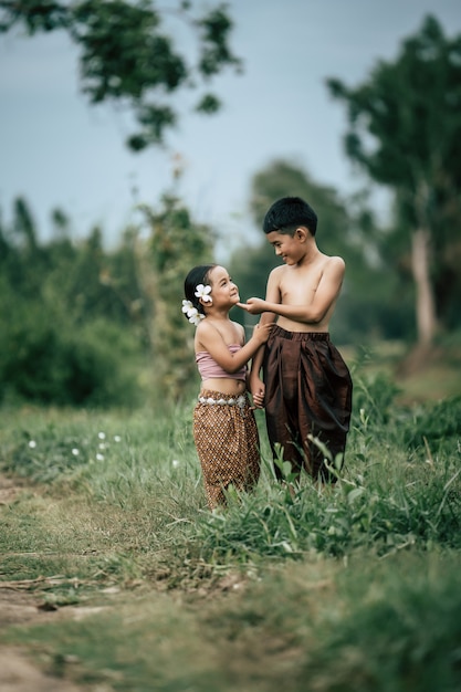 Retrato de un encantador niño asiático sin camisa y una niña en traje tradicional tailandés y poner una hermosa flor en su oreja, de pie de la mano y mirando al cielo con una sonrisa, espacio de copia