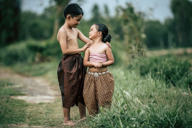 Foto gratuita retrato de un encantador niño asiático sin camisa y una niña en traje tradicional tailandés y poner una hermosa flor en su oreja, de pie de la mano y mirando al cielo con una sonrisa, espacio de copia