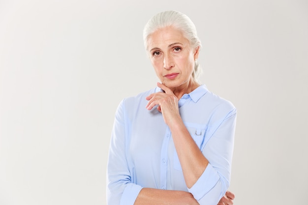 Retrato de encantadora anciana seria, en camisa azul, tocando su barbilla