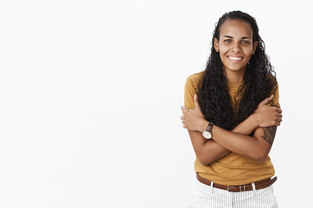 Retrato de una encantadora chica afroamericana amable y linda con el pelo largo y rizado temblando de frío como vistiendo una camiseta ligera en otoño sonriendo tontamente sobre la pared gris.