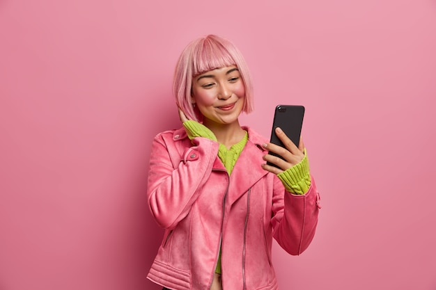 Retrato de encantadora chica asiática toma selfie en teléfono inteligente, hace una foto de sí misma para blog de moda, tiene peinado rosa, sonríe positivamente