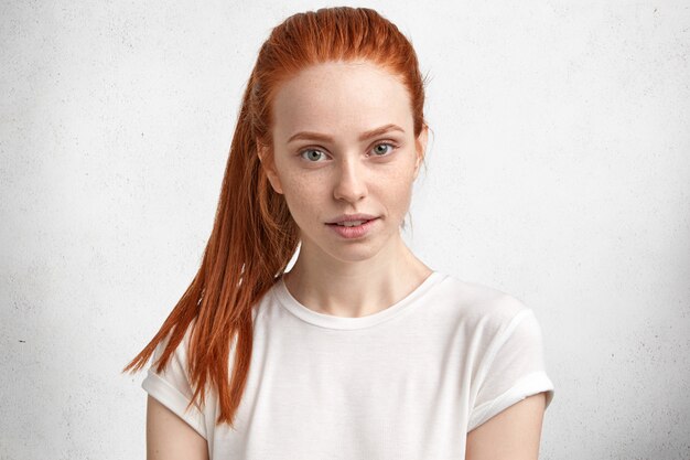 Retrato de encantadora hermosa estudiante con cola de caballo de jengibre, mira con confianza a la cámara, posa contra la pared de hormigón, piensa cómo pasar las vacaciones con placer.