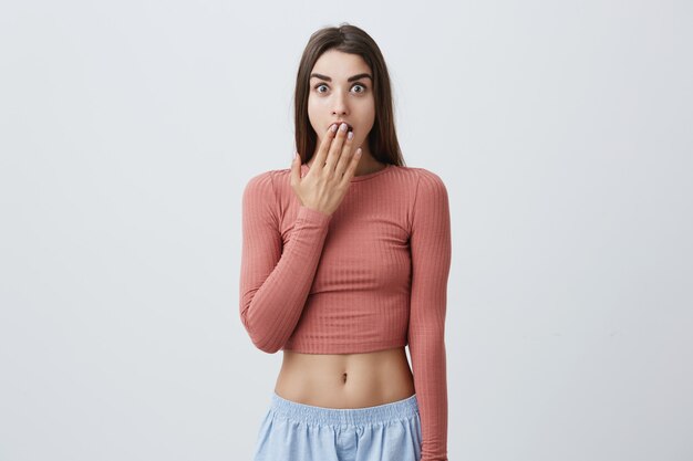 Retrato de encantadora joven morena caucásica divertida con el pelo largo en top rosa y jeans azul mirando con los ojos abiertos, cerrando la boca abierta con la mano