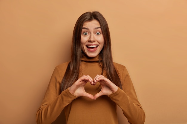 El retrato de la encantadora mujer alegre da forma a un gesto de corazón sobre el pecho, expresa amor y simpatía al novio, tiene una expresión divertida