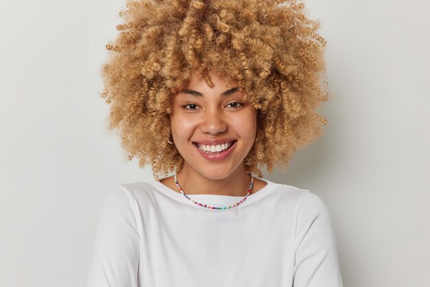 El retrato de una encantadora mujer feliz de cabello rizado sonríe con dientes y se ve con una expresión alegre que usa una camiseta informal y un collar que se mantiene positivo aislado sobre el fondo blanco Concepto de personas y emociones