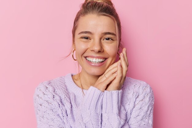 Retrato de una encantadora mujer joven que sonríe con dientes mantiene las manos cerca de la cara se ve contenta usa suéter de punto púrpura y aretes posa sobre fondo rosa Emociones felices y concepto de expresiones faciales