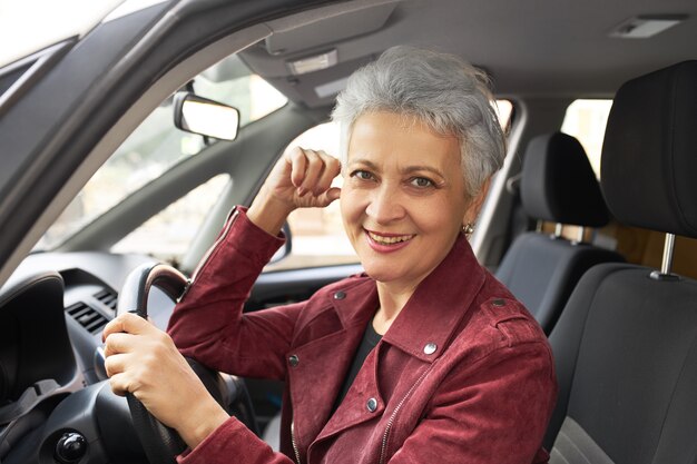 Retrato de encantadora mujer madura feliz con pelo gris corto sentado en el asiento del conductor