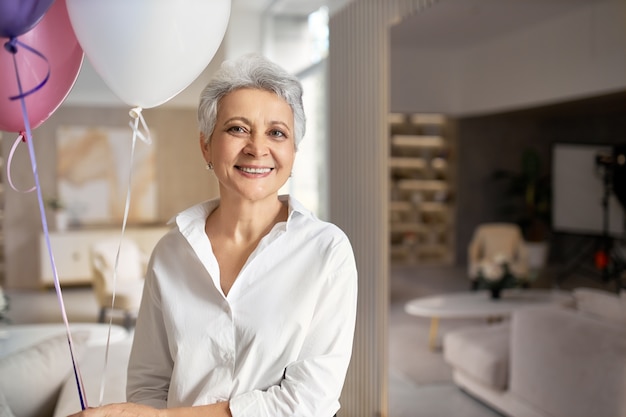 Retrato de encantadora mujer madura feliz vistiendo camisa formal divirtiéndose en su fiesta de jubilación, posando en el interior de la oficina con globos de helio