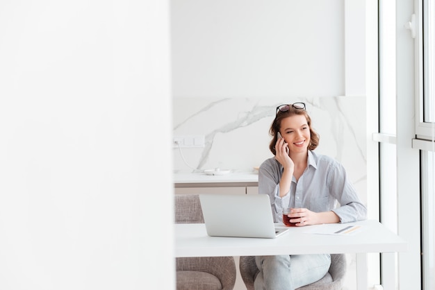 Foto gratuita retrato de encantadora mujer morena feliz hablando por teléfono móvil mientras está sentado y sosteniendo una taza de té caliente, en el interior