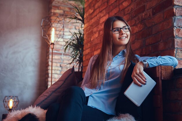 Retrato de una encantadora mujer de negocios morena con gafas y camisa azul sostiene una tableta y se sienta en un sofá en una habitación con diseño de loft.