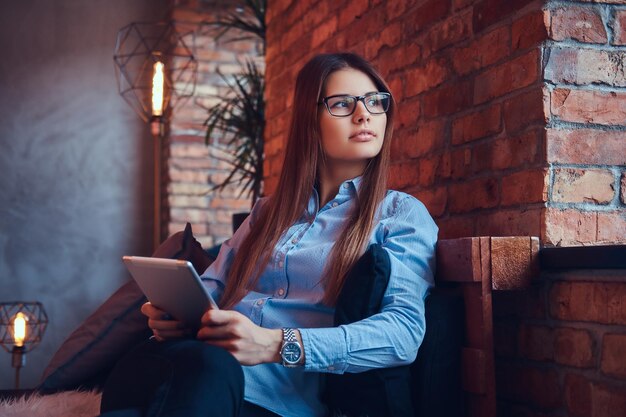 Retrato de una encantadora mujer de negocios morena con gafas y camisa azul sostiene una tableta y se sienta en un sofá en una habitación con diseño de loft.