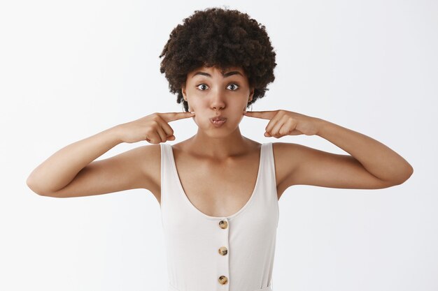 Retrato de encantadora mujer de piel oscura despreocupada y juguetona con peinado afro, haciendo pucheros, tocando las mejillas con los dedos índices y mirando con cara divertida