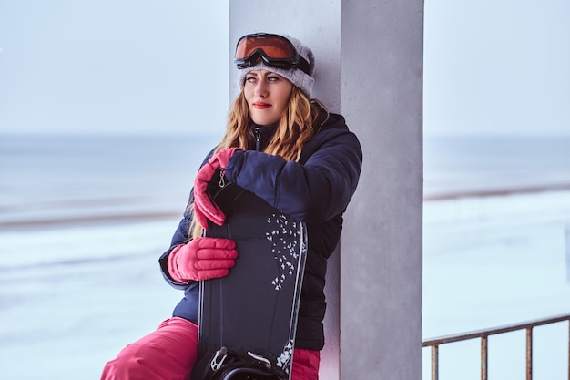 Retrato de una encantadora mujer rubia con sombrero de invierno y gafas sosteniendo una tabla de snowboard mientras se sienta en una barandilla cerca de la costa nevada del mar