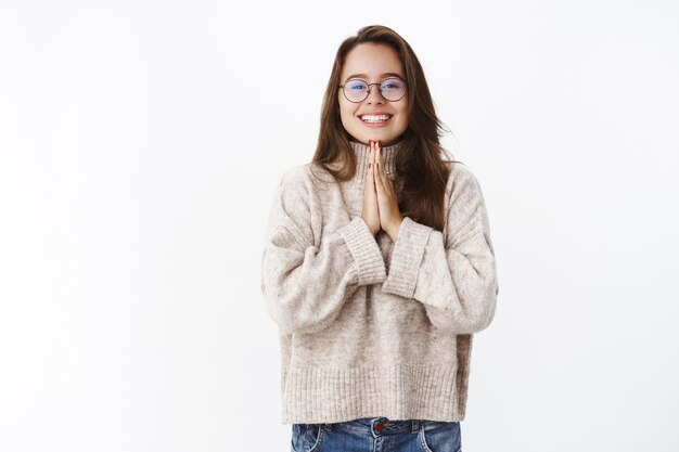 Retrato de encantadora niña coqueta tonta con gafas y un suéter tomados de la mano en rezar cerca del pecho y sonriendo ampliamente