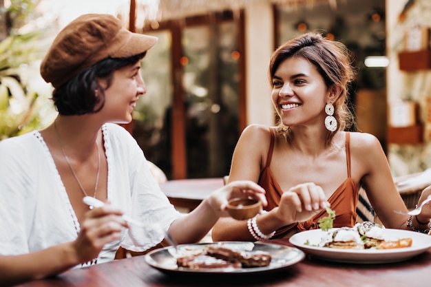 Retrato de encantadoras mujeres bronceadas de buen humor comiendo comida sabrosa en street cafe