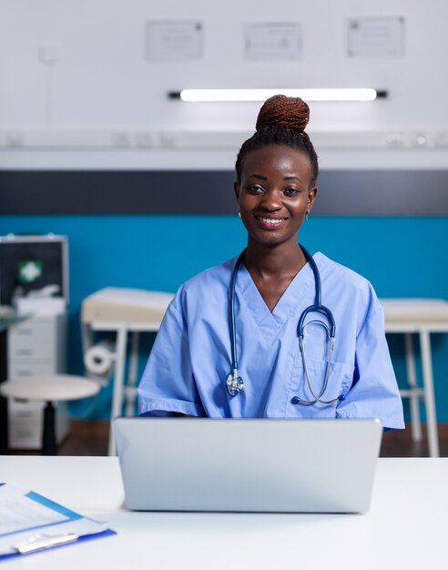 Retrato de enfermera afroamericana con ordenador portátil en el escritorio blanco