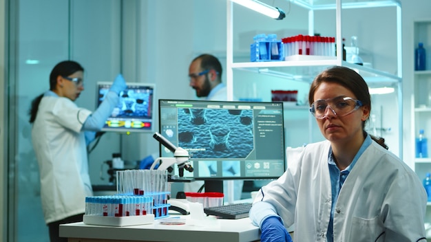 Retrato de enfermera científico mirando cansado a la cámara sentado en un moderno laboratorio equipado a altas horas de la noche. Equipo de especialistas que examinan la evolución del virus utilizando alta tecnología para la investigación y el desarrollo de vacunas.