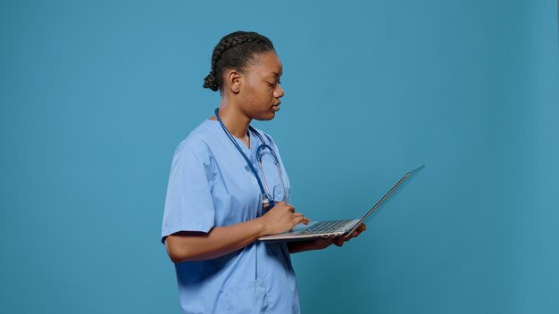 Retrato de una enfermera que trabaja en una laptop moderna en el estudio. Especialista en atención médica que sostiene una computadora con tecnología, usa un dispositivo para obtener experiencia médica. Asistente con estetoscopio