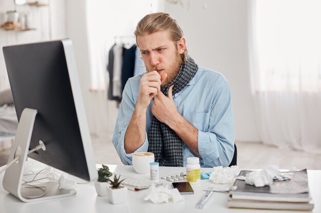 Retrato de enfermo enfermo barbudo gerente masculino tose, tiene resfriado y gripe. El joven rubio tiene nariz respingona, tos y resfriado, se sienta en el lugar de trabajo frente a la pantalla de la computadora. Enfermedad e infección