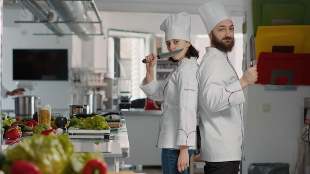 Retrato del equipo de chefs actuando divertido con cuchillos en la cocina del restaurante, haciendo bromas sobre el servicio de cocina profesional. Hombre y mujer alegres trabajando en un plato de comida gourmet con receta culinaria.