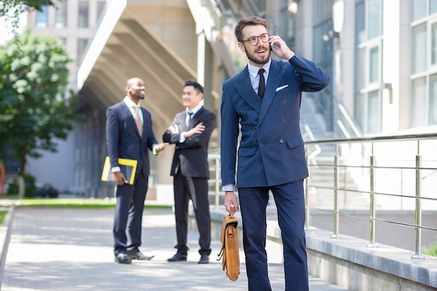 Retrato de equipo multiétnico de negocios. Tres hombres de pie contra el fondo de la ciudad. El primer plano de un hombre europeo hablando por teléfono. Otros hombres son chinos y afroamericanos.