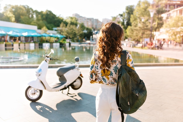 Retrato de espalda de chica delgada con cabello castaño rizado de pie junto a scooter con mochila de cuero