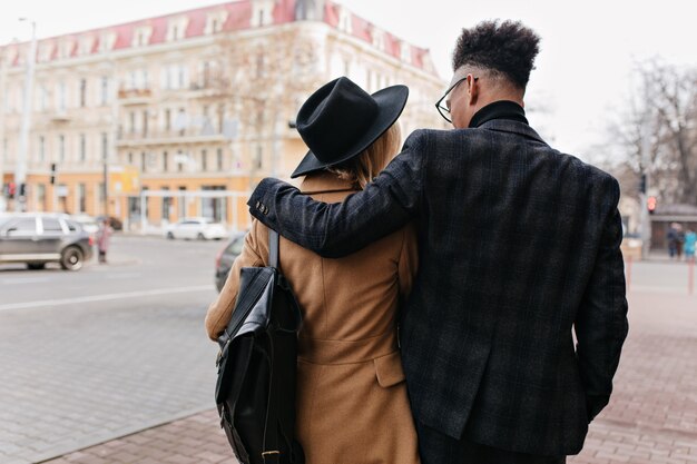 Retrato de espalda del hombre africano en traje elegante caminando por la ciudad con su novia. Chico rizado negro abrazando a mujer encantadora con sombrero y abrigo beige.