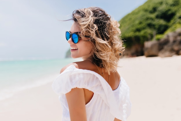 Retrato de espalda de mujer bronceada interesada escalofriante en el resort. Disparo al aire libre de adorable mujer con cabello ondulado ligero caminando por la playa.