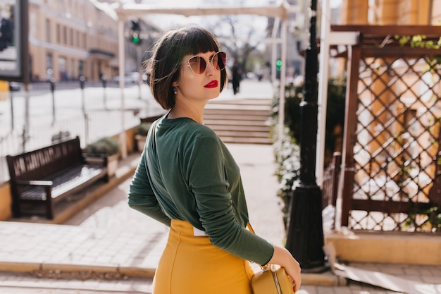 Retrato de espalda de mujer morena sensual con labios rojos mirando por encima del hombro. Retrato al aire libre de elegante dama europea en suéter verde caminando por la calle.