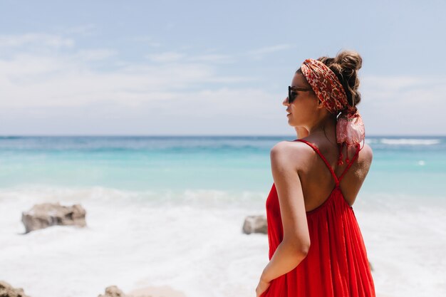 Retrato de espalda de niña despreocupada con piel bronceada mirando al horizonte. Foto de feliz modelo de mujer caucásica en traje rojo escalofriante en la costa del océano y disfrutando de la vista.