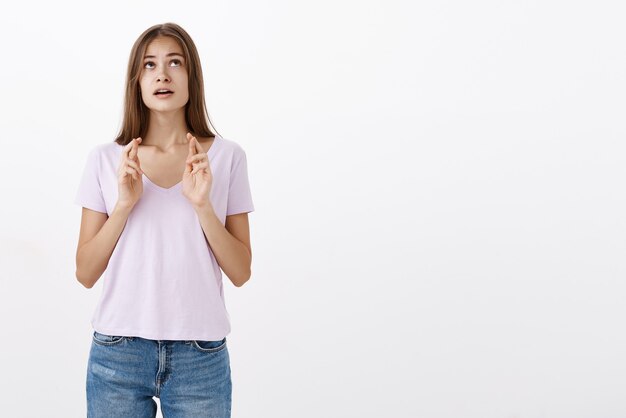 Retrato de esperanza, determinada y preocupada linda hembra europea con cabello castaño en traje casual abriendo la boca mirando hacia arriba preocupado y cruzando los dedos para la buena suerte