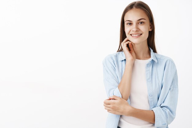 Retrato de esperanza tierna y femenina morena europea en blusa azul sobre camiseta blanca tocando la cara suavemente y sonriendo