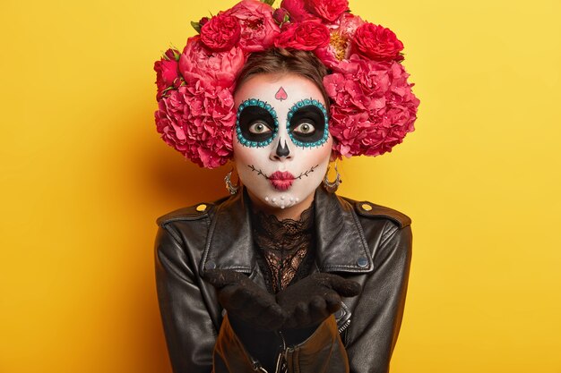 Retrato de esqueleto femenino apuesto se prepara para el carnaval mexicano, sopla beso al aire, usa maquillaje de calavera, vestido con chaqueta negra, tiene aspecto aterrador, aislado sobre fondo amarillo. Pintura corporal y arte facial