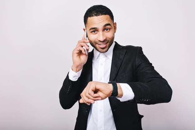 Retrato con estilo elegante joven con camisa blanca y chaqueta negra hablando por teléfono, mostrando el reloj y sonriendo. Alegre, hombre de negocios, reunión, sonreír, trabajo