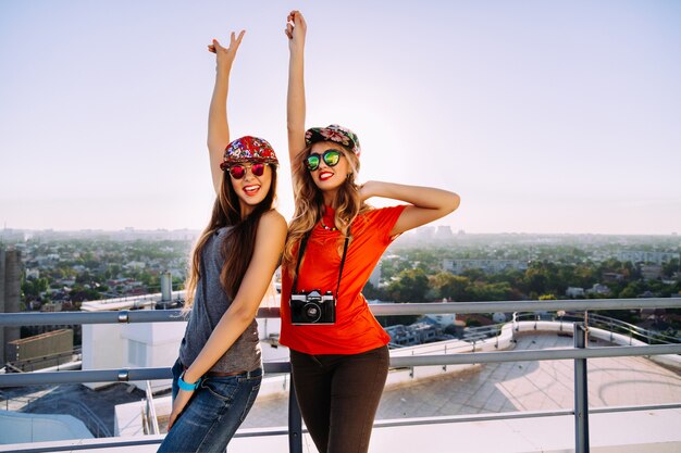 Retrato de estilo de vida al aire libre de dos mejores amigos bastante elegantes posando en el techo con una vista increíble de la ciudad, levantan las manos gritando riendo volviéndose locos y disfrutando de su libertad