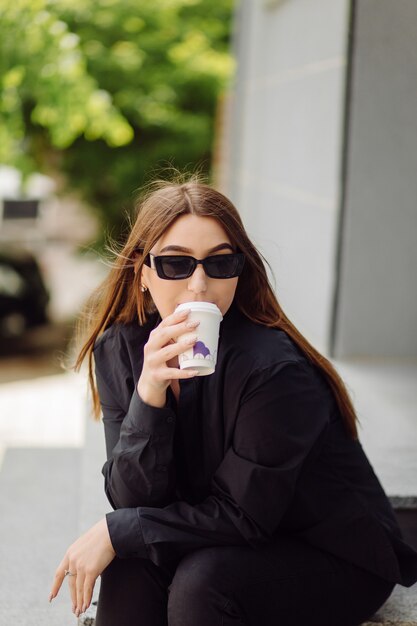 Retrato de estilo de vida al aire libre de la impresionante chica morena. Tomando café y caminando por las calles de la ciudad.